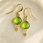 Peridot Ballet Drops: a pair of faceted green gemstone earrings with gold hooks and small dangling gold accents. AI-styled closeup; original product photographs remain in the gallery.