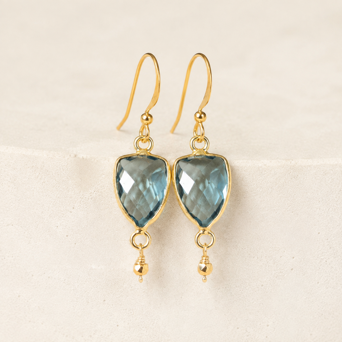 The Isla Drops in London Blue Quartz — refined close-up of faceted stones and gold-plated silver hooks on ivory stone.