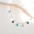 The Azzurra Necklace in gold, with seven blue gemstone drops on a fine chain, shown against pale paper.
