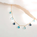 The Azzurra Necklace in gold, with seven blue gemstone drops on a fine chain, shown against pale paper.