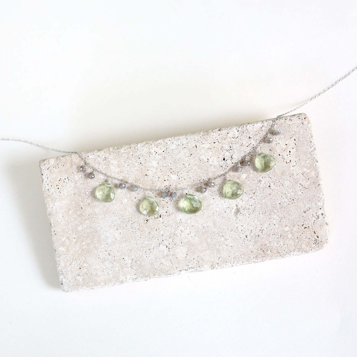 The Prasiolite Necklace with pale green gemstone drops and silver-tone chain, shown on a stone display.