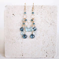 The Cascade Drops in gold, with cascading blue gemstones and open circular links, shown on pale stone.