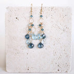 The Cascade Drops in gold, with cascading blue gemstones and open circular links, shown on pale stone.