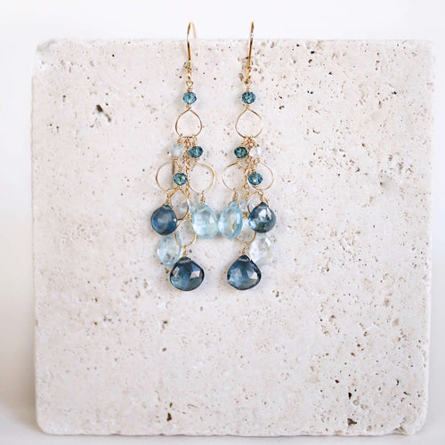 The Cascade Drops in gold, with cascading blue gemstones and open circular links, shown on pale stone.