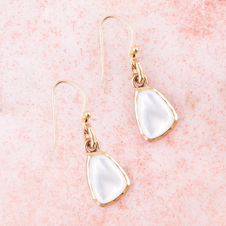 The Nacre Drops laid diagonally on a pink surface, showing white mother-of-pearl faces and bronze settings.