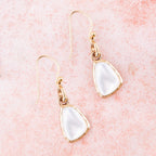 The Nacre Drops laid diagonally on a pink surface, showing white mother-of-pearl faces and bronze settings.